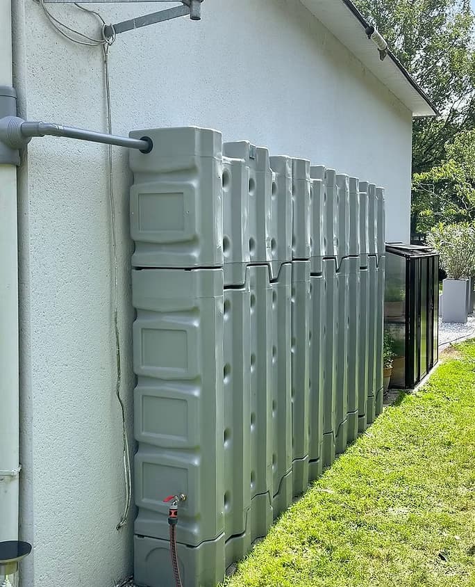 Récupérateur d'eau de pluie aérien Murdeau coloris gris pierre installé contre un mur
